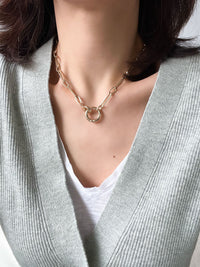 Gold paperclip chain necklace with elongated rectangular links and a circular carabiner lock pendant in the center.