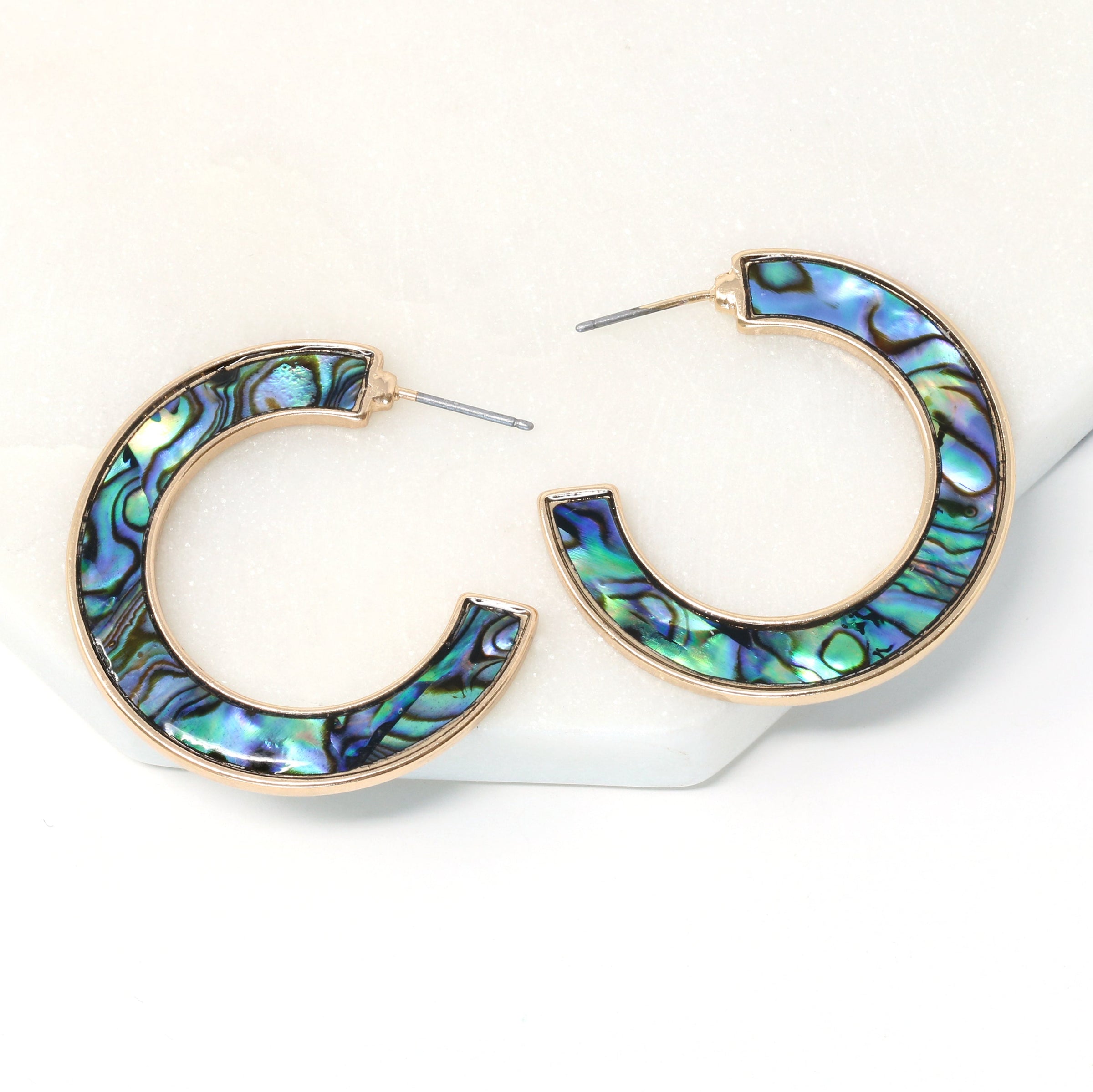 A pair of metal-mounted natural abalone shell hoop earrings. The iridescent abalone shells display beautiful shades of blue, green, black and purple, with a unique natural pattern visible from both sides.