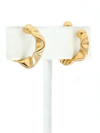 A pair of gold cuff hoop earrings with a twisted, organic wavy design, displayed on a white jewelry stand against a white background.