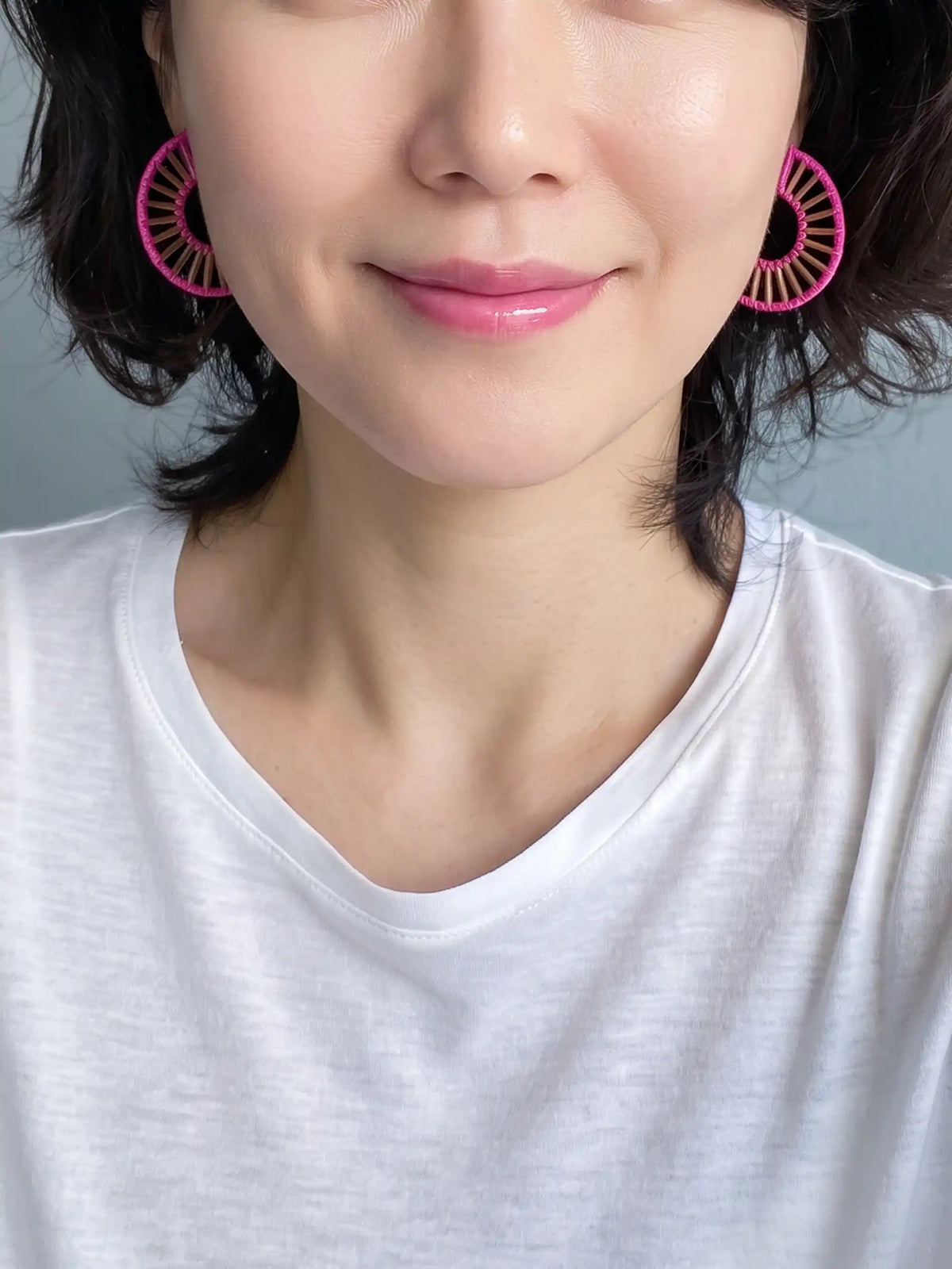 A close-up of a woman wearing bright pink circular fan-shaped earrings and a white V-neck t-shirt against a soft gray background.