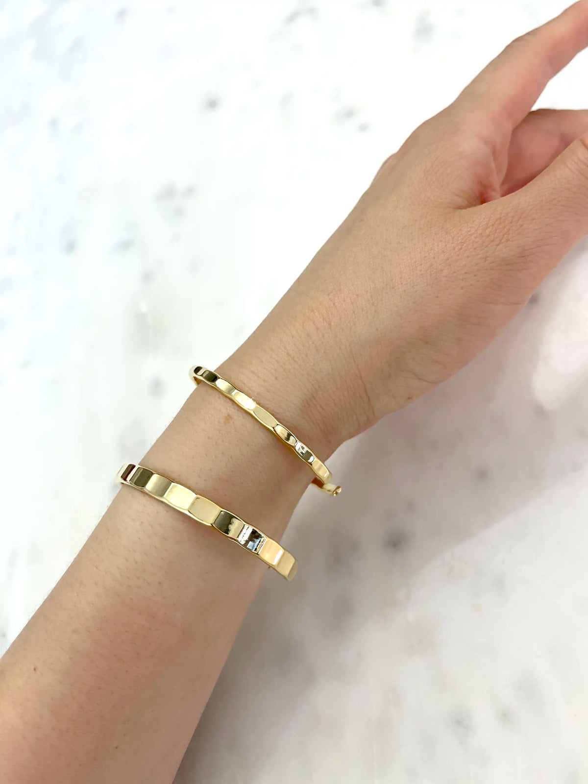 Two slim gold bangle bracelets with small clear crystal accents, shown worn on a person's wrist against a light marble background.