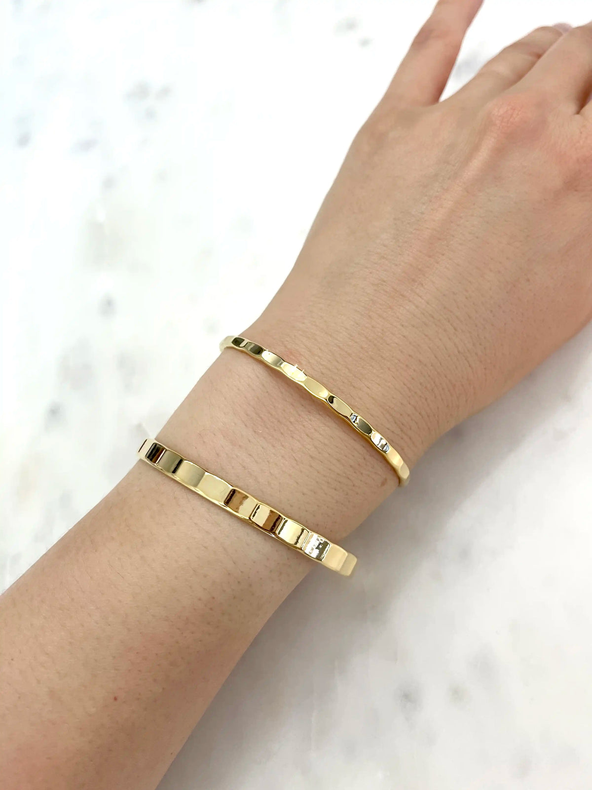 Two slim gold bangle bracelets with small clear crystal accents, shown worn on a person's wrist against a light marble background.