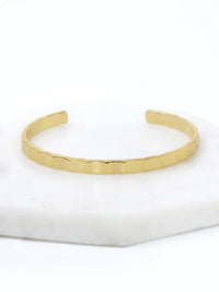 A hammered gold-tone cuff bracelet resting on a white marble surface.