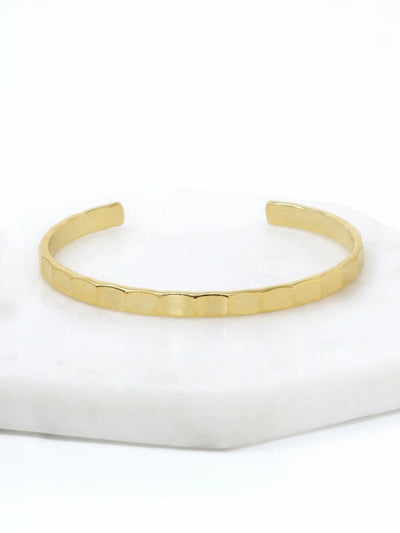 A hammered gold-tone cuff bracelet resting on a white marble surface.