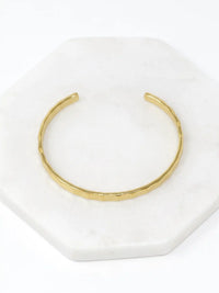 A hammered gold-tone cuff bracelet resting on a white marble surface.