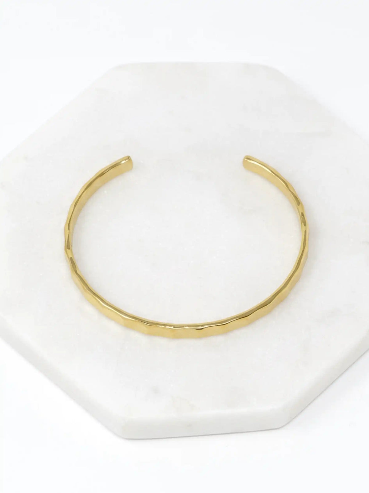 A hammered gold-tone cuff bracelet resting on a white marble surface.