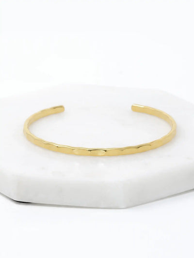 A hammered gold-tone cuff bracelet resting on a white marble surface.