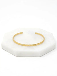  A hammered gold-tone cuff bracelet resting on a white marble surface.