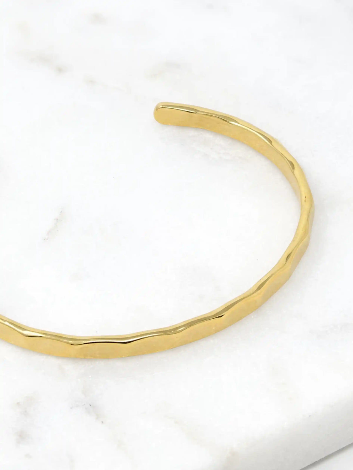A hammered gold-tone cuff bracelet resting on a white marble surface.