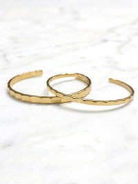 An open-ended, polished gold-tone metal cuff bracelet with a unique hammered or faceted texture along the main body. The lighting highlights the texture and smooth ends.