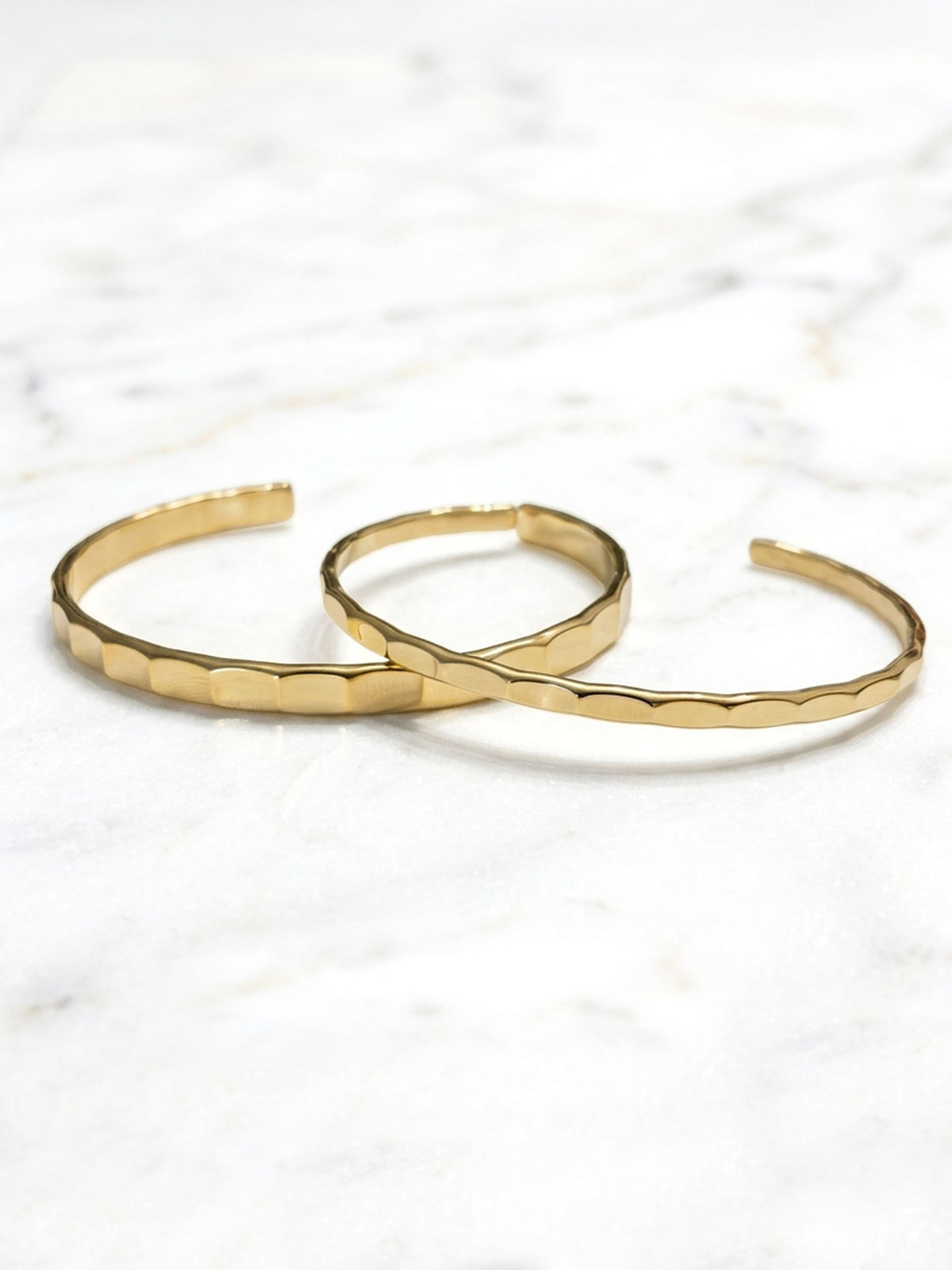 An open-ended, polished gold-tone metal cuff bracelet with a unique hammered or faceted texture along the main body. The lighting highlights the texture and smooth ends.