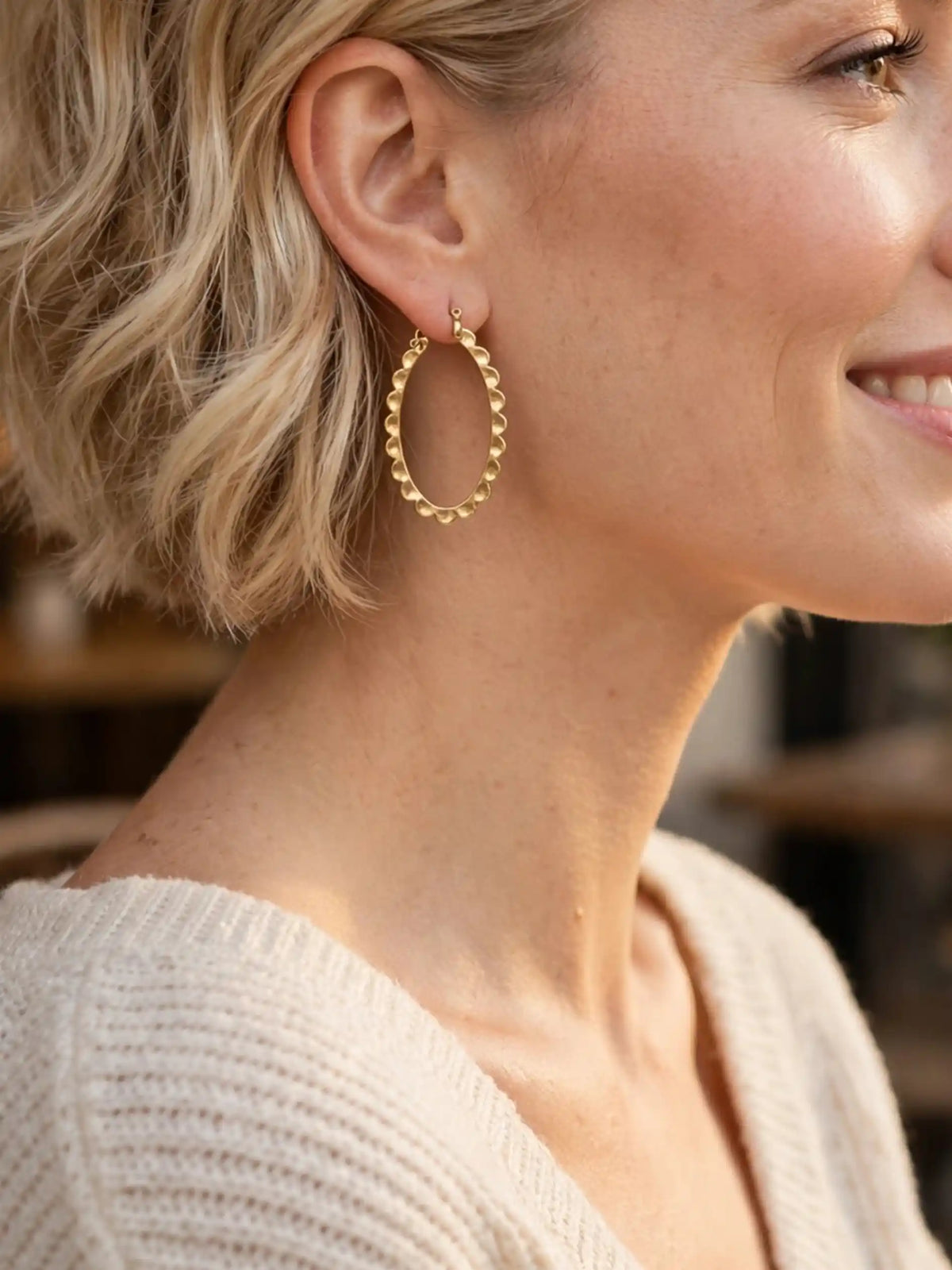 A close-up profile of a smiling woman with short blonde hair wearing a gold, scalloped oval hoop earring and a cream-colored knit sweater.