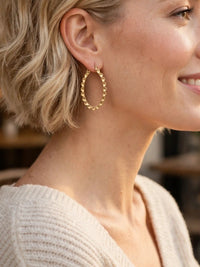A close-up profile of a smiling woman with short blonde hair wearing a gold, scalloped oval hoop earring and a cream-colored knit sweater.
