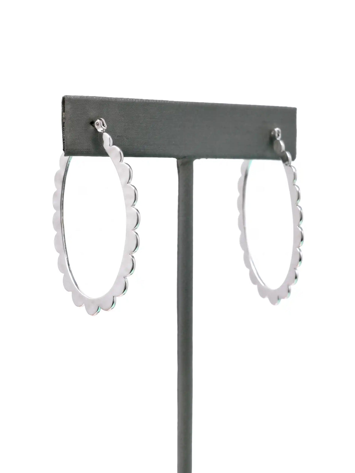 A pair of silver-colored hoop earrings with a scalloped edge design. The earrings are displayed on a gray stand against a white background. The hoops are oval-shaped and have a shiny finish.