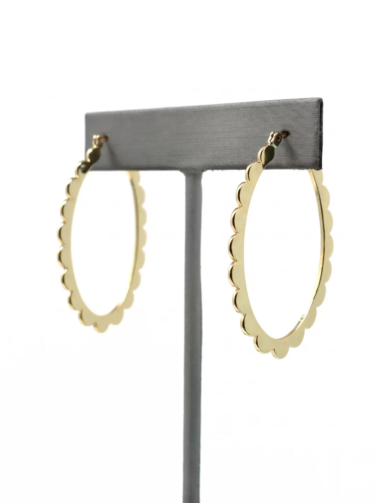 A pair of gold-colored hoop earrings with a scalloped edge design. The earrings are displayed on a gray stand against a white background. The hoops are oval-shaped and have a shiny finish.