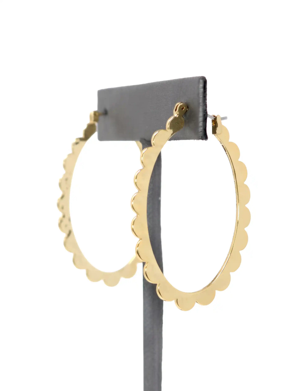 A pair of gold-colored hoop earrings with a scalloped edge design. The earrings are displayed on a gray stand against a white background. The hoops are oval-shaped and have a shiny finish.