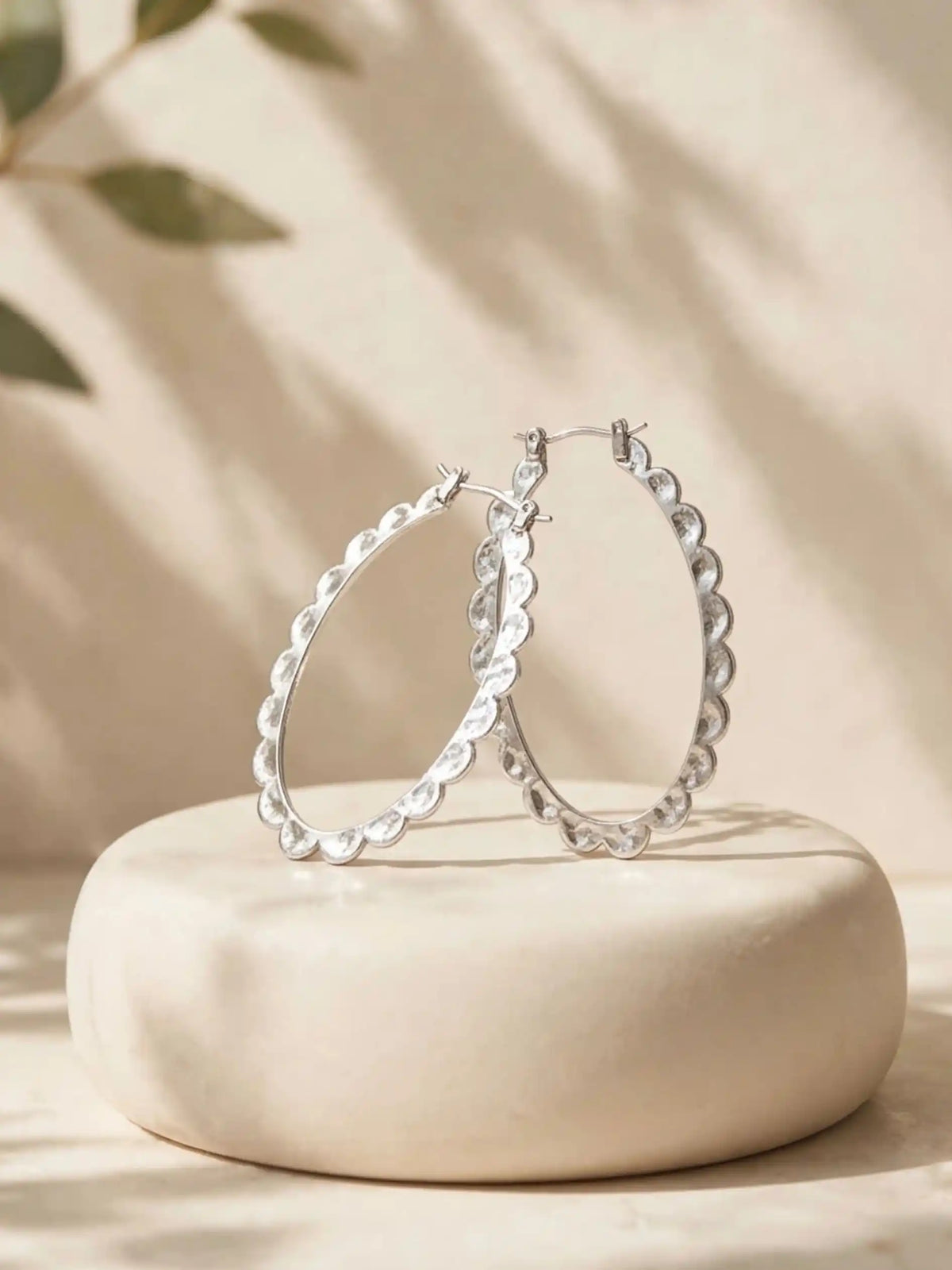 A pair of silver scalloped oval hoop earrings standing upright on a smooth, round beige stone display base, illuminated by warm sunlight with soft leafy shadows cast in the background.