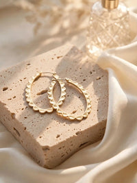 A pair of gold, scalloped oval hoop earrings resting on a textured light beige stone block, surrounded by softly draped cream-colored fabric in warm sunlight, with a blurred faceted glass bottle in the background.