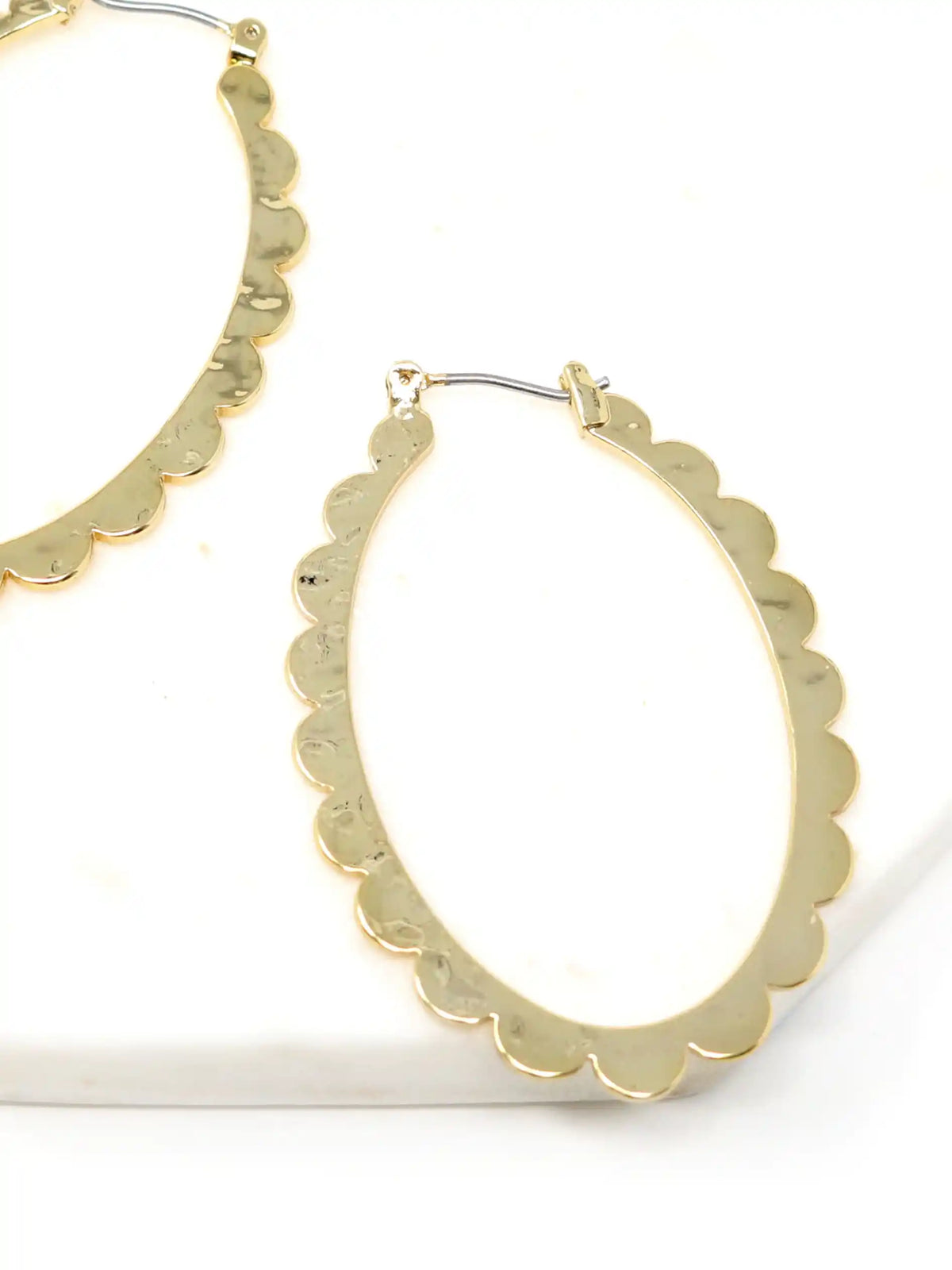 A pair of gold-colored hoop earrings with a scalloped edge design. The earrings are displayed on a gray stand against a white background. The hoops are oval-shaped and have a shiny finish.