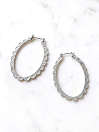 Two silver-colored oval hoop earrings with a scalloped edge design. The earrings are displayed on a white surface. The earrings have a simple hook closure.