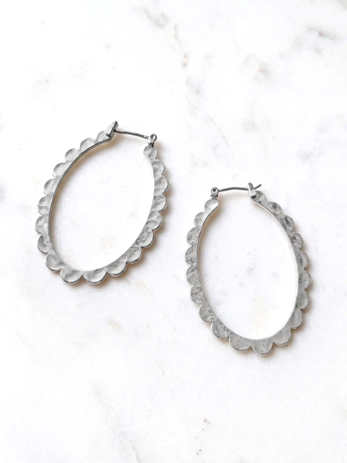 Two silver-colored oval hoop earrings with a scalloped edge design. The earrings are displayed on a white surface. The earrings have a simple hook closure.