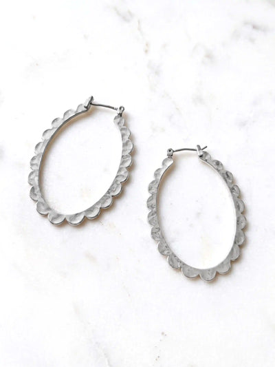 Two silver-colored oval hoop earrings with a scalloped edge design. The earrings are displayed on a white surface. The earrings have a simple hook closure.