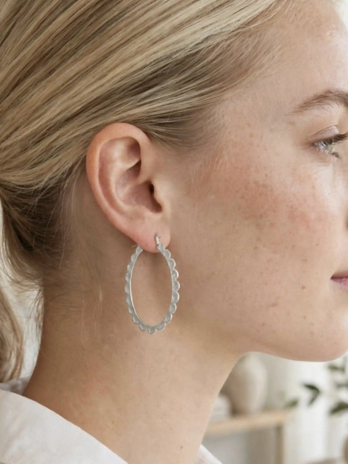 A silver hoop earring with a scalloped edge is worn on the ear. The person has blonde hair and is wearing a white top. The earring is the focal point of the image.