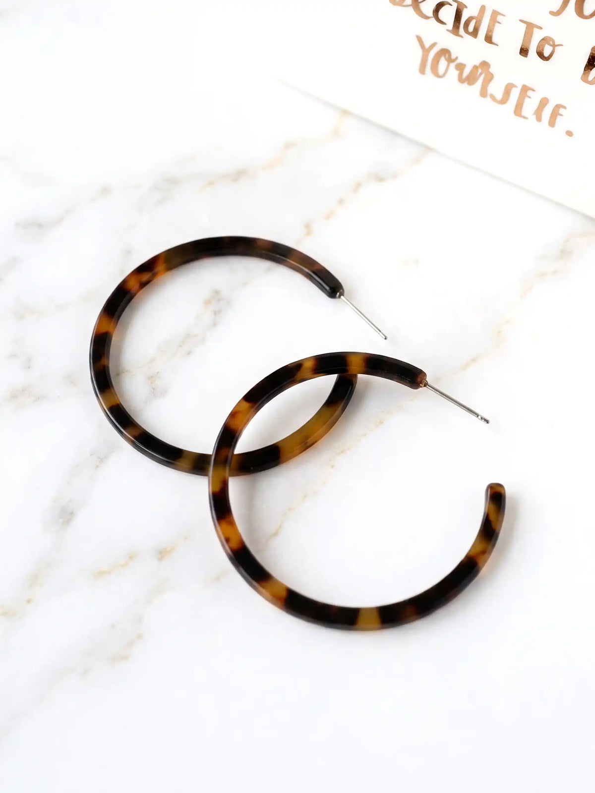 A pair of 50mm brown tortoiseshell resin hoop earrings resting flat on a sleek white marble surface with soft natural lighting and subtle shadows.