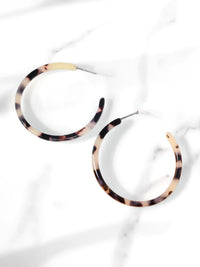 A pair of 50mm light brown tortoiseshell resin hoop earrings resting flat on a sleek white marble surface with soft natural lighting and subtle shadows.