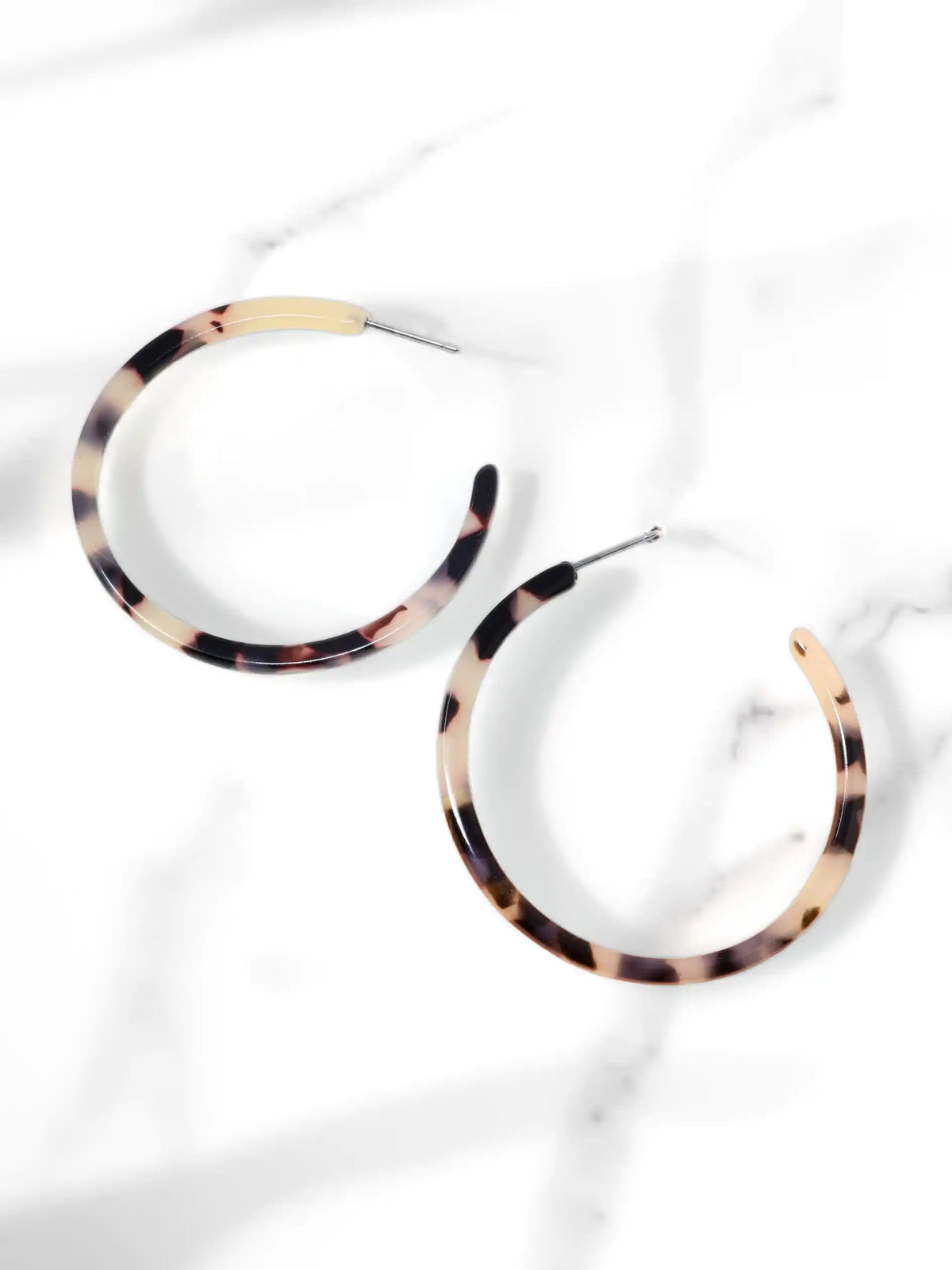 A pair of 50mm light brown tortoiseshell resin hoop earrings resting flat on a sleek white marble surface with soft natural lighting and subtle shadows.