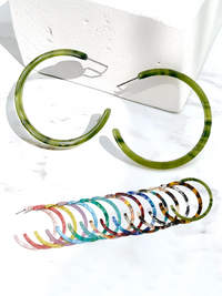 Two large open-ended hoop earrings with a green marbled tortoiseshell pattern are displayed on a white marble surface next to a white textured block, with a row of smaller multicolored tortoiseshell hoop earrings fanned out below.
