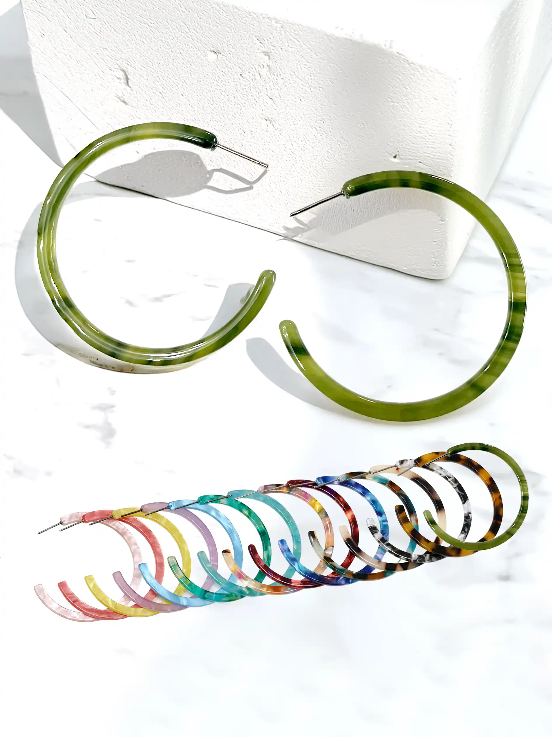 Two large open-ended hoop earrings with a green marbled tortoiseshell pattern are displayed on a white marble surface next to a white textured block, with a row of smaller multicolored tortoiseshell hoop earrings fanned out below.