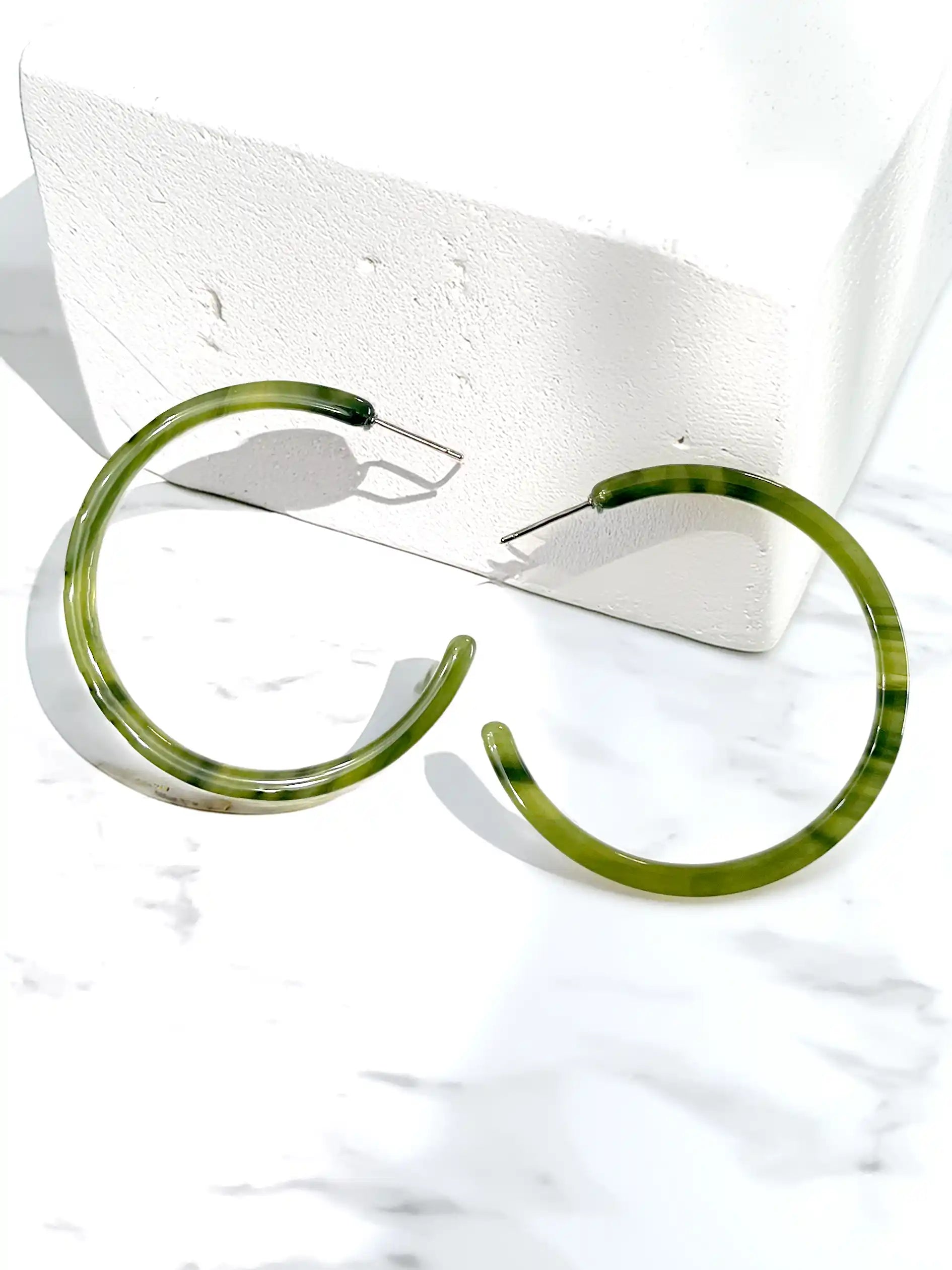 A pair of thick, open-ended hoop earrings with a marbled olive green and black pattern, displayed on a white marble surface next to a white textured block.