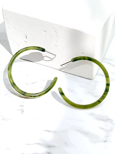 A pair of thick, open-ended hoop earrings with a marbled olive green and black pattern, displayed on a white marble surface next to a white textured block.