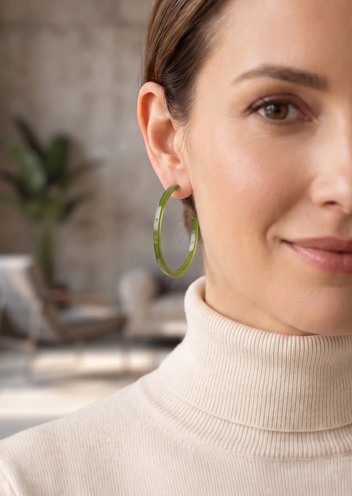 A close-up of a woman wearing a single large translucent olive green hoop earring, paired with a cream ribbed turtleneck sweater in a softly blurred indoor setting.
