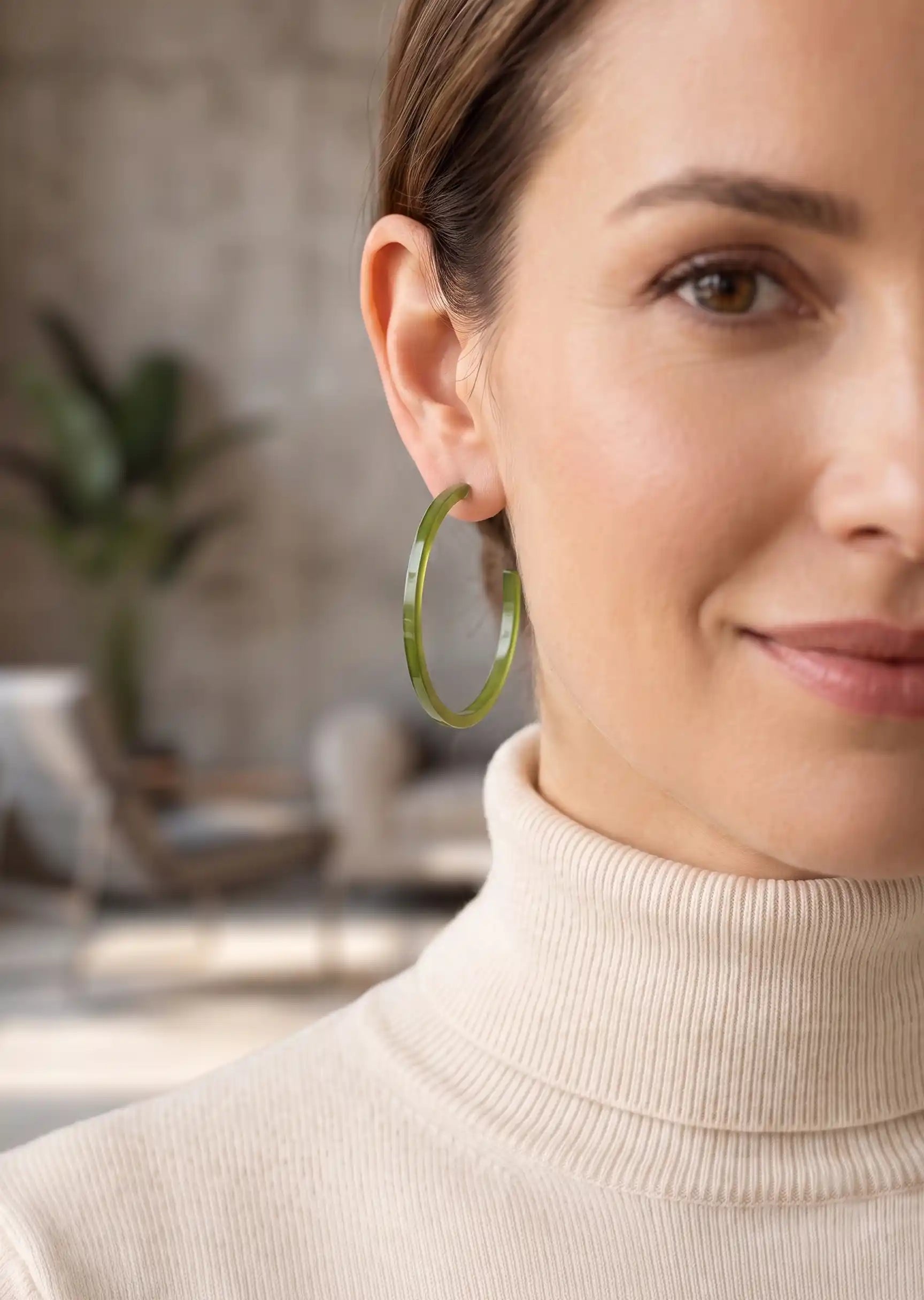 A close-up of a woman wearing a single large translucent olive green hoop earring, paired with a cream ribbed turtleneck sweater in a softly blurred indoor setting.