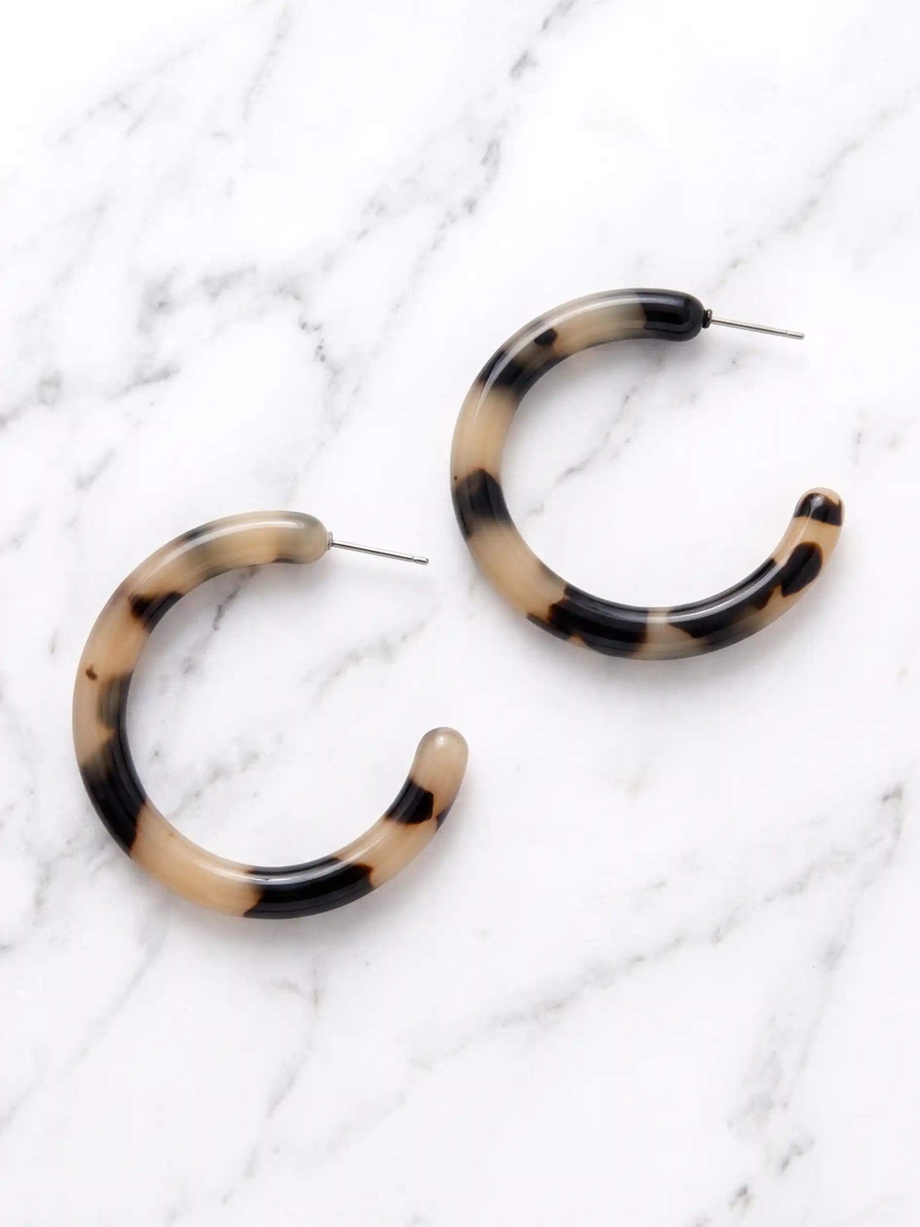 Light brown tortoise resin hoop earrings with a marbled pattern of beige, brown, and black, displayed on a white marble background.