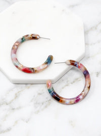 Marbled hoop earrings with a multicolor pattern of pink, blue, purple, and brown, displayed on a white hexagonal marble tray over a white marble surface.