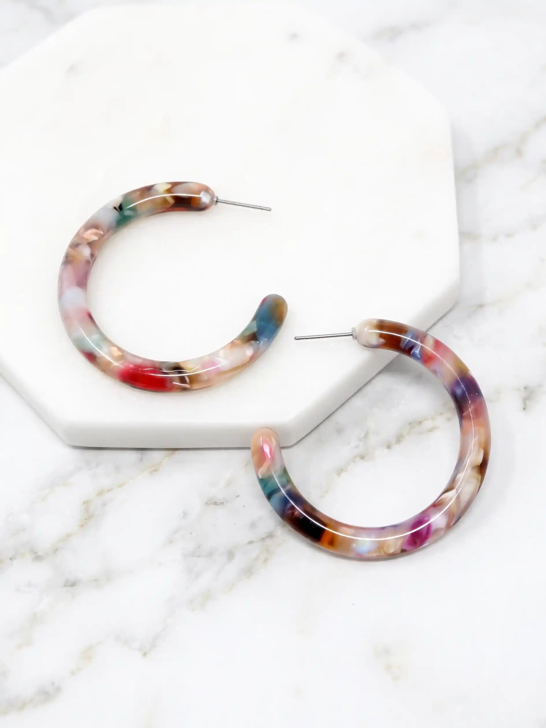 Marbled hoop earrings with a multicolor pattern of pink, blue, purple, and brown, displayed on a white hexagonal marble tray over a white marble surface.