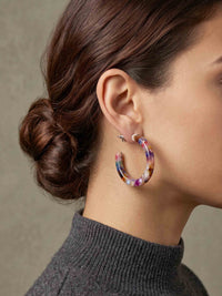 A close-up of a woman's ear and side profile wearing a large marbled hoop earring in purple, pink, and blue tones, paired with a dark gray ribbed turtleneck sweater against a neutral background.