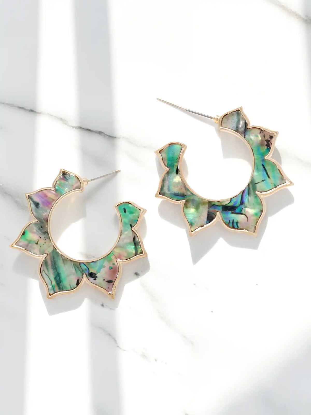 A pair of gold floral hoop earrings with green iridescent abalone shell inlay, displayed on a white marble surface with soft shadows.