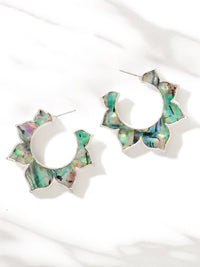 A pair of silver floral hoop earrings with irregular, petal-like edges inlaid with iridescent green, blue, and pink abalone shell, displayed on a white marble surface.
