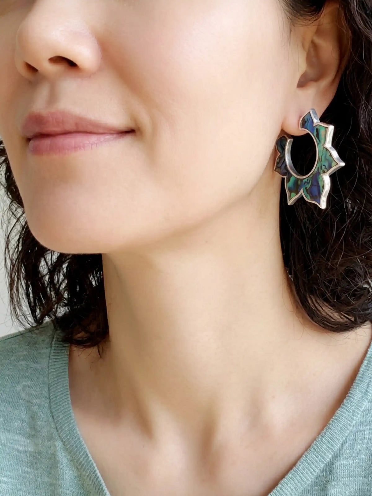 A close-up of a model wearing a silver-toned floral earring with iridescent blue and green inlay.