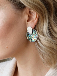 A close-up image of woman wearing a silver-tone round disc stud earrings with colorful iridescent abalone shell inlay in blue, green, and pink.