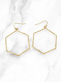 A pair of gold-tone hexagonal hoop earrings with a textured, hammered finish and fishhook backings, displayed on a white marble background.