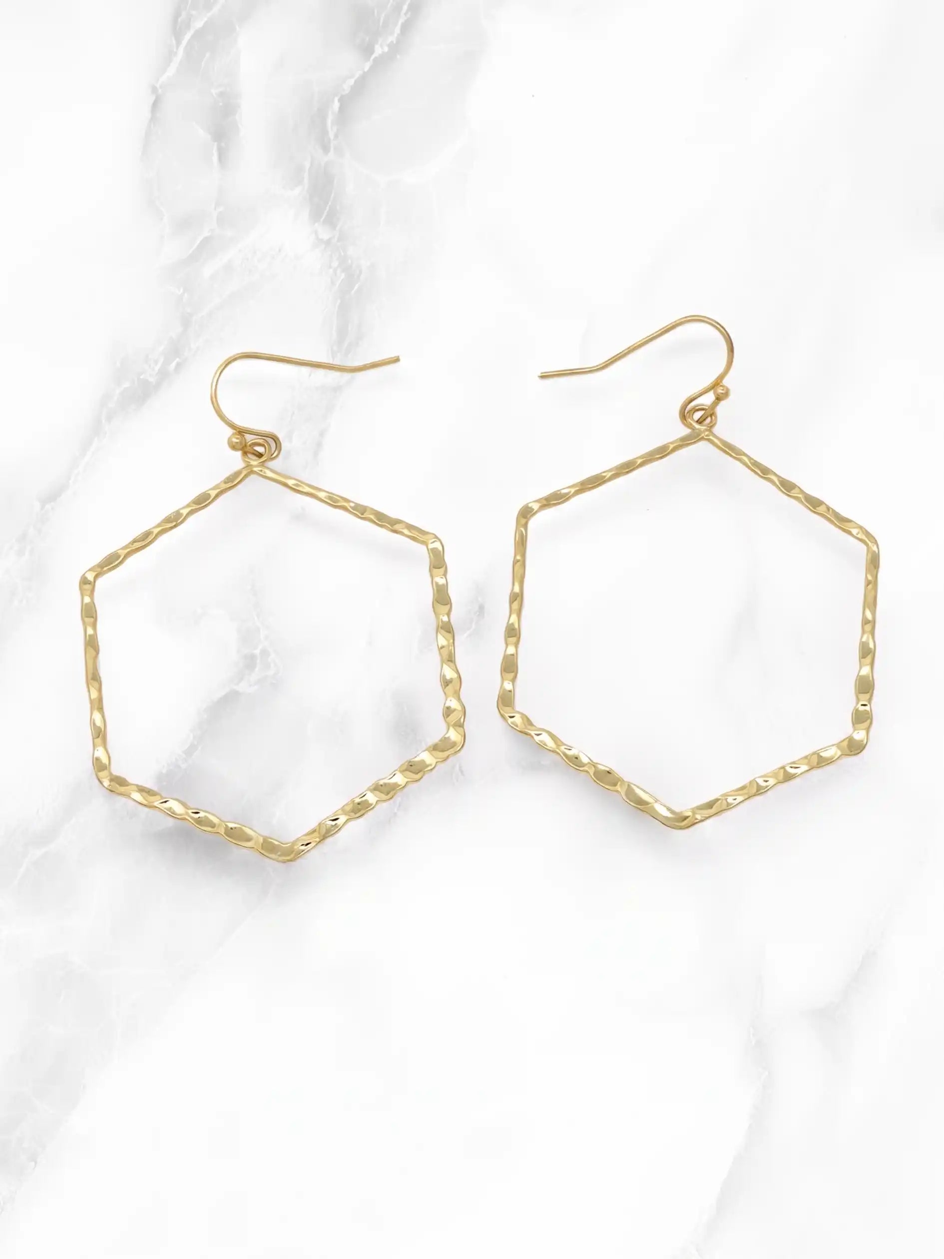 A pair of gold-tone hexagonal hoop earrings with a textured, hammered finish and fishhook backings, displayed on a white marble background.