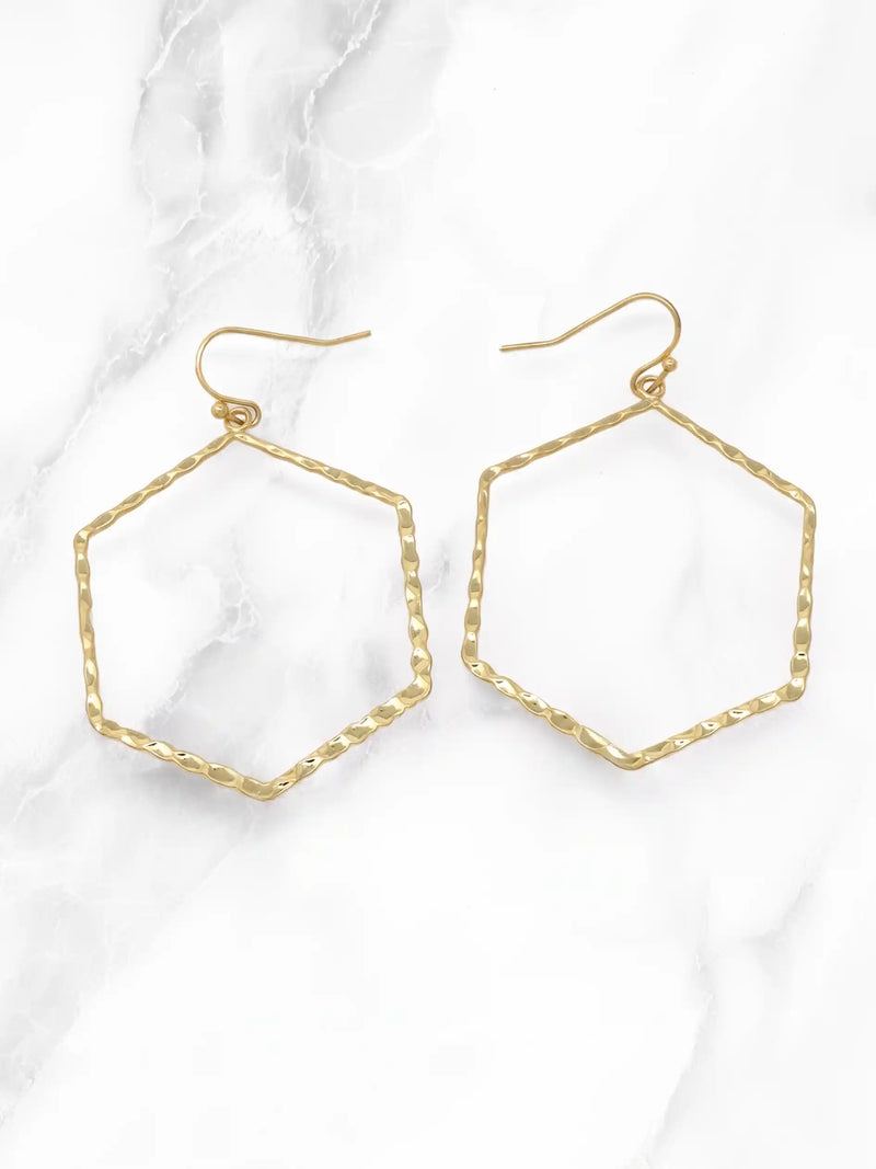 A pair of gold-tone hexagonal hoop earrings with a textured, hammered finish and fishhook backings, displayed on a white marble background.