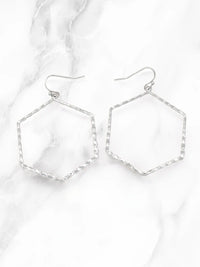 A pair of silver-tone geometric drop earrings with textured hexagonal wire frames and fishhook backings, displayed on a white marble surface.