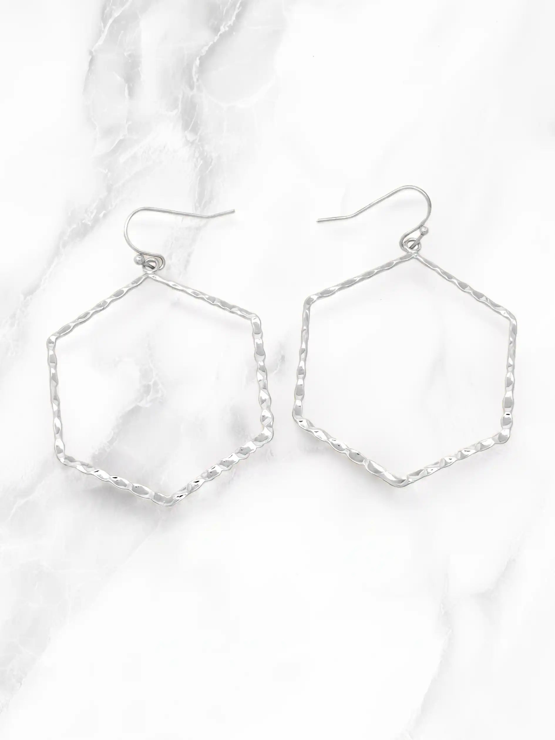 A pair of silver-tone geometric drop earrings with textured hexagonal wire frames and fishhook backings, displayed on a white marble surface.