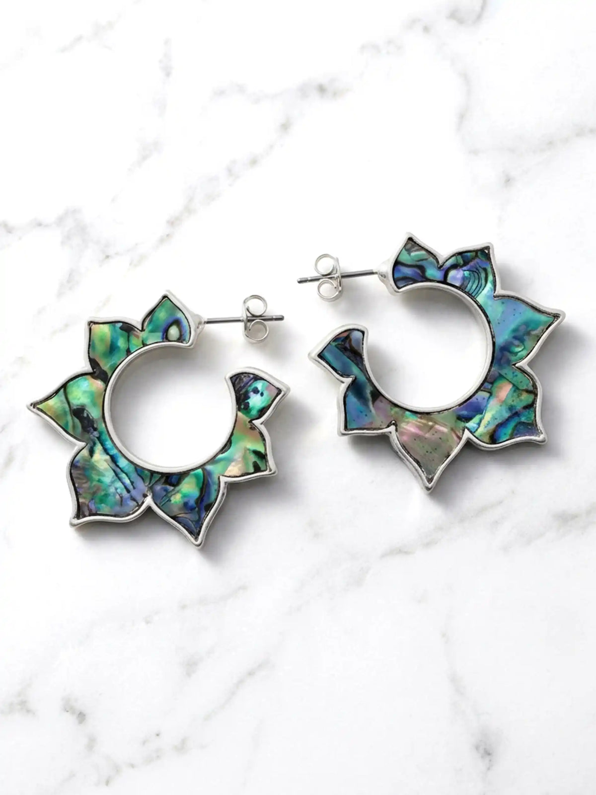 Silver floral hoop earrings with layered abalone shell inlays in shades of green, blue, and purple, displayed on a white marble background.